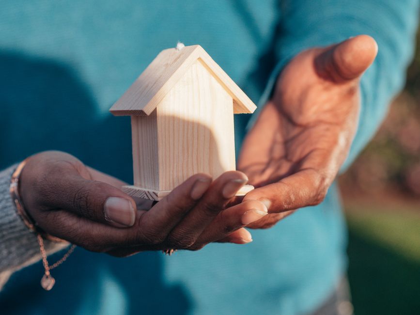 In a brightly lit, minimalist setting, a pair of hands are gently cupped around a small model house and a piggy bank, illustrating the concept of protecting home equity and savings.