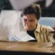 Person with a concerned expression sits at a clean, modern desk, reviewing a stack of official-looking papers and using a ...