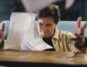 Person with a concerned expression sits at a clean, modern desk, reviewing a stack of official-looking papers and using a ...