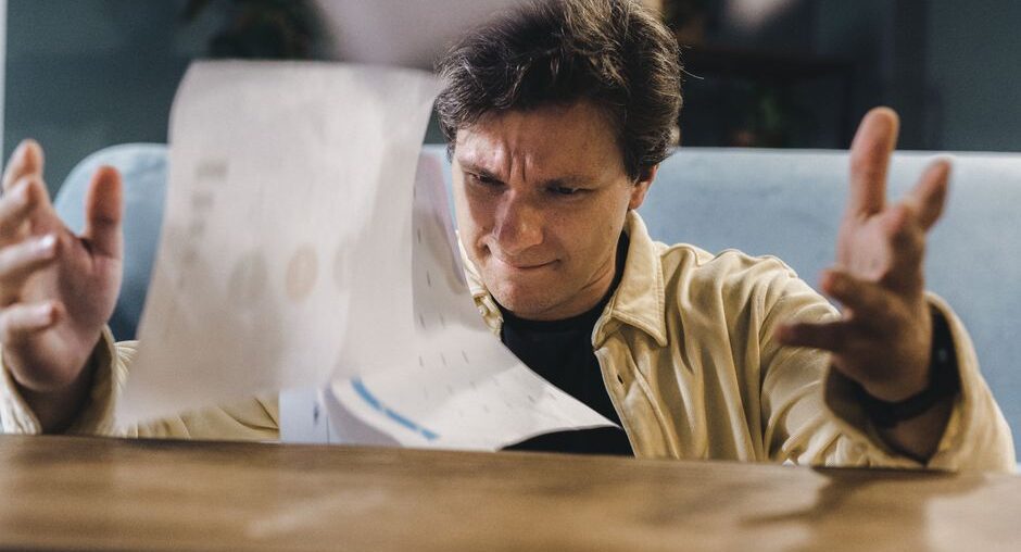 Person with a concerned expression sits at a clean, modern desk, reviewing a stack of official-looking papers and using a ...
