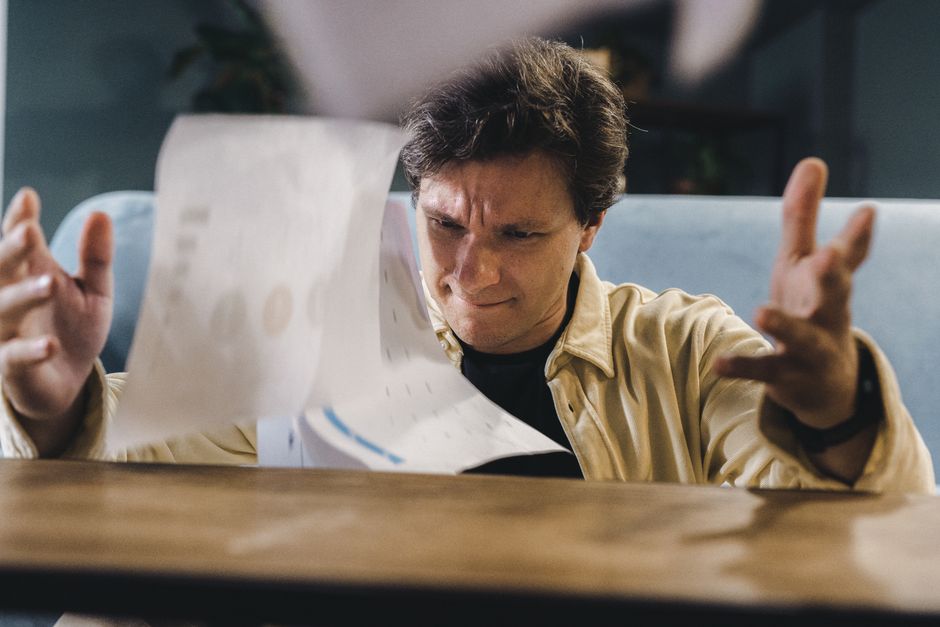 Person with a concerned expression sits at a clean, modern desk, reviewing a stack of official-looking papers and using a ...